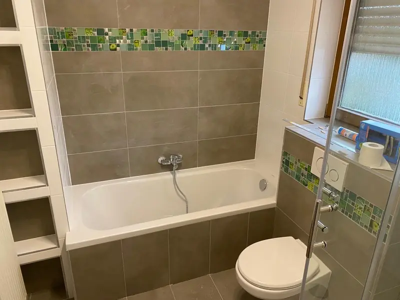 Renoviertes Badezimmer mit Badewanne, beigen Fliesen und grüner Mosaik-Bordüre